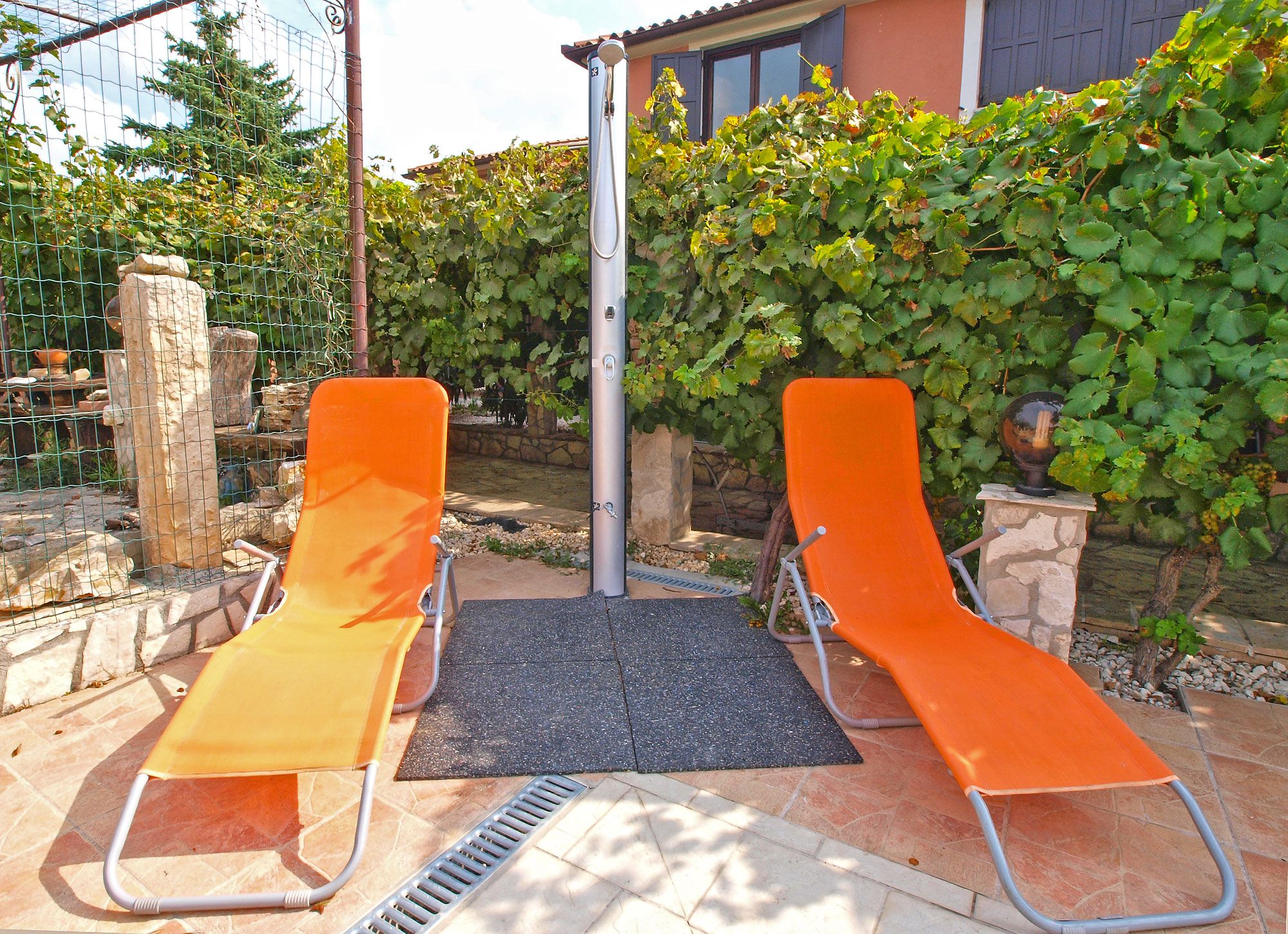 Two orange lounge chairs with a metal frame, a black mat, and a metal pole in a garden area with a stone wall and greenery.