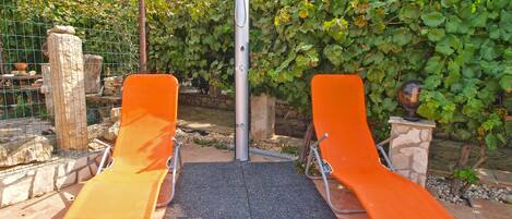 Two orange lounge chairs with a metal frame, a black mat, and a metal pole in a garden area with a stone wall and greenery.