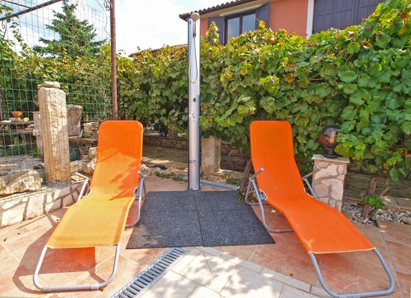 Two orange lounge chairs with a metal frame, a black mat, and a metal pole in a garden area with a stone wall and greenery.