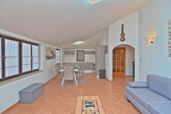 A spacious living area with a sofa, a dining table with chairs, a kitchen in the background, and a large window with blinds.
