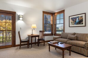 A living room with a sofa, a coffee table, a lamp, and a chair. There are large windows with blinds, a wall-mounted light fixture, and a framed picture on the wall.