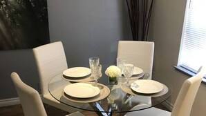 A dining area with a glass table surrounded by four white chairs, a vase with white flowers, and a window with blinds. - La Cassita Beautiful Condo WOW!!!! (Las Vegas)