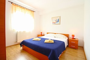 A bedroom with a bed, two bedside tables, a window with curtains, a radiator, and a painting on the wall.