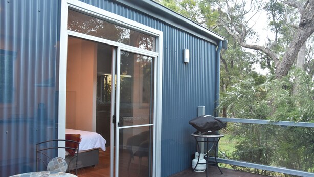 A balcony with a table and chairs, a grill, and a sliding glass door leading to a bedroom with a bed and a chair. - Manuka Retreat, Free unlimited WIFI.... (Halls Gap)