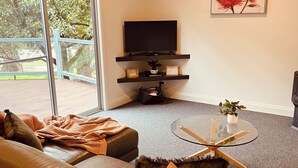 A living room with a leather sofa, a glass coffee table, a flat-screen TV, and a large window with a view of trees. - Manuka Retreat, Free unlimited WIFI.... (Halls Gap)