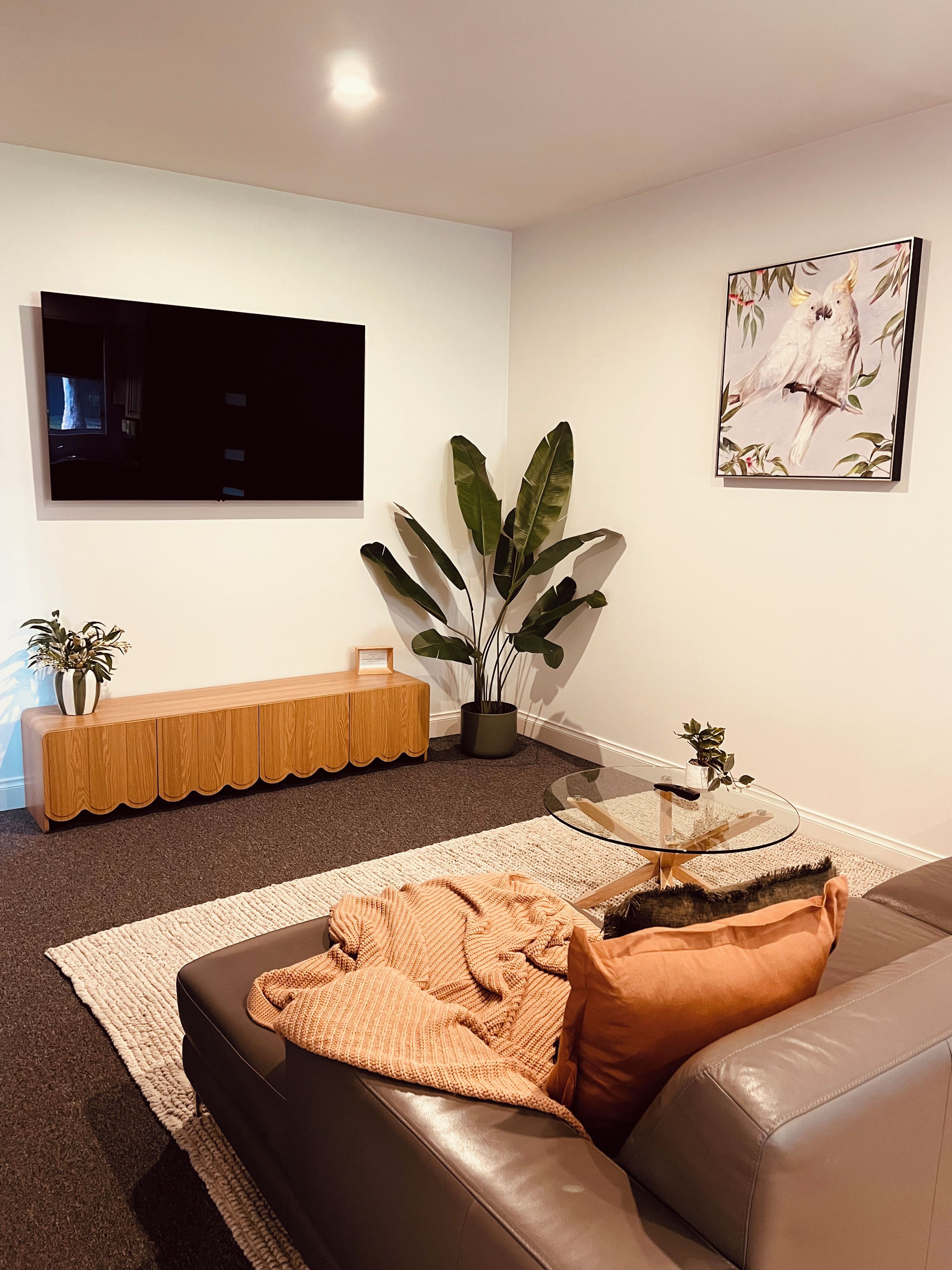 Ett modernt vardagsrum med en soffa i läder, ett soffbord i glas, en stor krukväxt, ett avlastningsbord i trä, en platt-TV som är monterad på väggen och ett inramat konstverk.