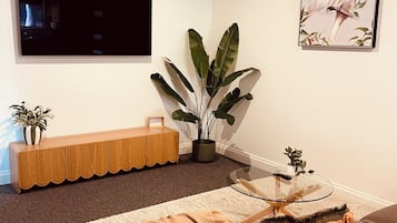 Ett modernt vardagsrum med en soffa i läder, ett soffbord i glas, en stor krukväxt, ett avlastningsbord i trä, en platt-TV som är monterad på väggen och ett inramat konstverk.