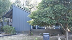 A modern building with a sloped roof and large windows, surrounded by a garden with a tree and a stone planter. - Manuka Retreat, Free unlimited WIFI.... (Halls Gap)
