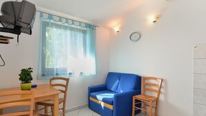 Een kamer met een blauwe bank, houten stoelen, een eettafel, een potplant, een raam met dunne gordijnen en een wandklok.