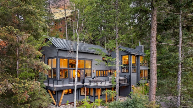Une cabane à deux étages dotée de grandes fenêtres, d’une terrasse et de fondations en pierre, le tout entouré d’arbres.