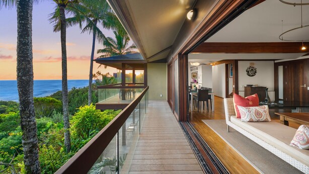 Un balcon avec vue sur l’océan et les palmiers, un coin salon avec un canapé et des coussins, et un espace repas avec une table et des chaises.