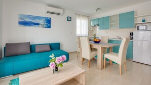 A modern living room with a teal sofa, a dining table with chairs, a small coffee table, and a kitchen area with teal cabinets and white appliances. - Apartment 813/1901 (Istria - Medulin), 90m from the beach (Medulin)