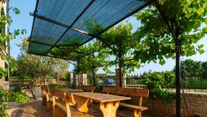 An outdoor patio with wooden benches and a table, covered by a blue mesh canopy, surrounded by greenery and a stone wall. - Apartment 813/1902 (Istria - Medulin), 90m from the beach (Medulin)
