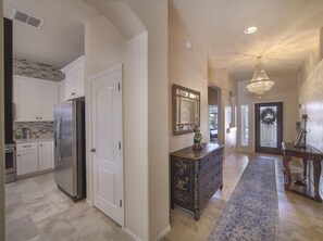 A spacious kitchen with white cabinets, a stainless steel refrigerator, and a stone backsplash. A hallway with a patterned rug leads to a dining area with a chandelier.