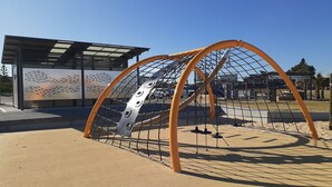 A playground with a climbing structure and a building with a patterned facade in the background. - Absolute Waterfront Anna Bay (Anna Bay)