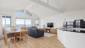 A modern kitchen with a black countertop, white cabinets, and a large island. There is a dining area with a wooden table and chairs, and a grey sofa. The room has large windows with a view of the ocean. - Absolute Waterfront Anna Bay (Anna Bay)