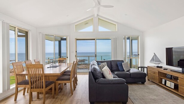 A spacious living room with a large TV, a dining area with wooden furniture, and a view of the ocean through large windows. - Absolute Waterfront Anna Bay (Anna Bay)