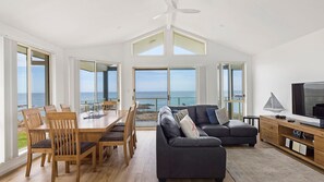 A spacious living room with a large TV, a dining area with wooden furniture, and a view of the ocean through large windows. - Absolute Waterfront Anna Bay (Anna Bay)