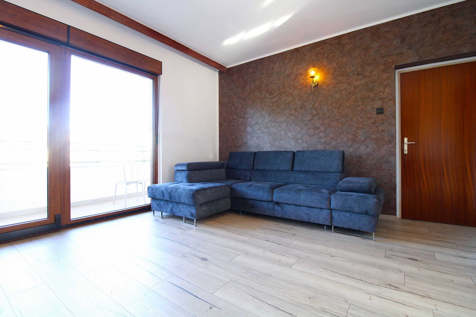 Una sala de estar moderna con un sofá seccional, piso de madera, un amplio ventanal corredero y una pared con textura.