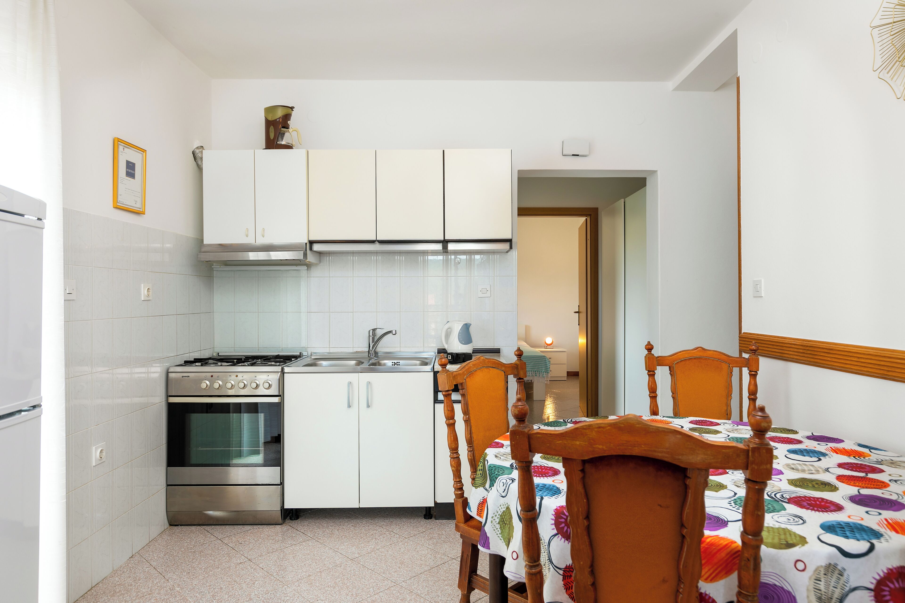 A kitchen with white cabinets, a stove, and a sink. There is a wooden chair and a table with a colorful tablecloth.
