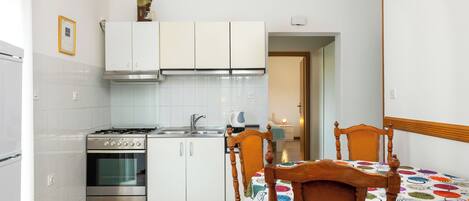 A kitchen with white cabinets, a stove, and a sink. There is a wooden chair and a table with a colorful tablecloth.