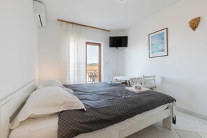 A bedroom with a bed, a TV mounted on the wall, a desk with a chair, and a window with curtains.