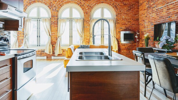 A kitchen with a brick wall, a large island with a sink, and a dining area with a table and chairs. - Authentic loft for family of 4 in the heart of Old Quebec (La Cité-Limoilou)