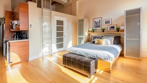 A bedroom with a large bed, a wooden bench, a wardrobe, and a kitchenette. - Authentic loft for family of 4 in the heart of Old Quebec (La Cité-Limoilou)