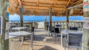An outdoor dining area with wooden tables and chairs, a thatched roof, and a view of the beach and ocean. - Ocean Views in Palmetto Dunes- Great amenities! (Hilton Head)