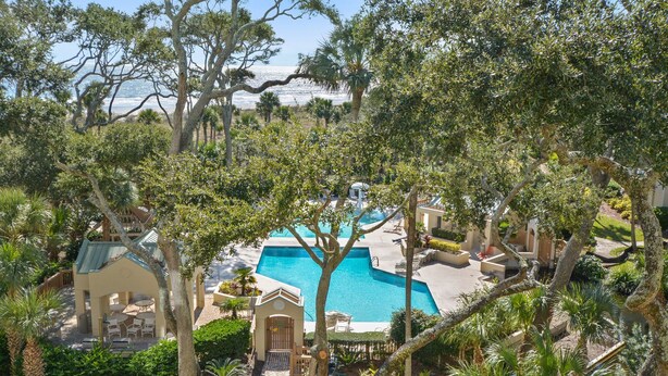 A resort with a central swimming pool, surrounded by lush greenery and palm trees. - Ocean Views in Palmetto Dunes- Great amenities! (Hilton Head)