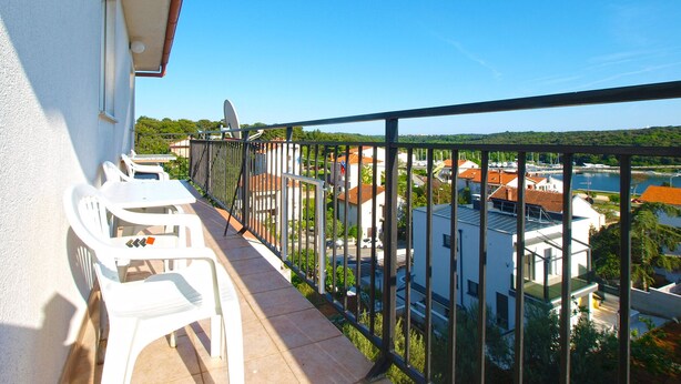 A balcony with white plastic chairs and a table, overlooking a residential area with houses and a body of water.