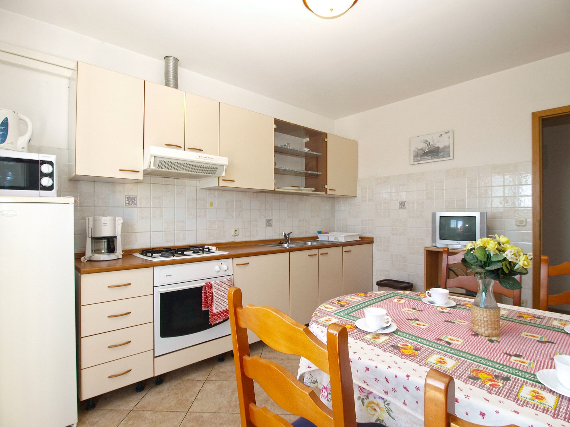 Eine kompakte Küche mit Herd, Mikrowelle und Kaffeemaschine. Ein Esstisch mit Stühlen und ein kleiner Fernseher auf einem Schrank.
