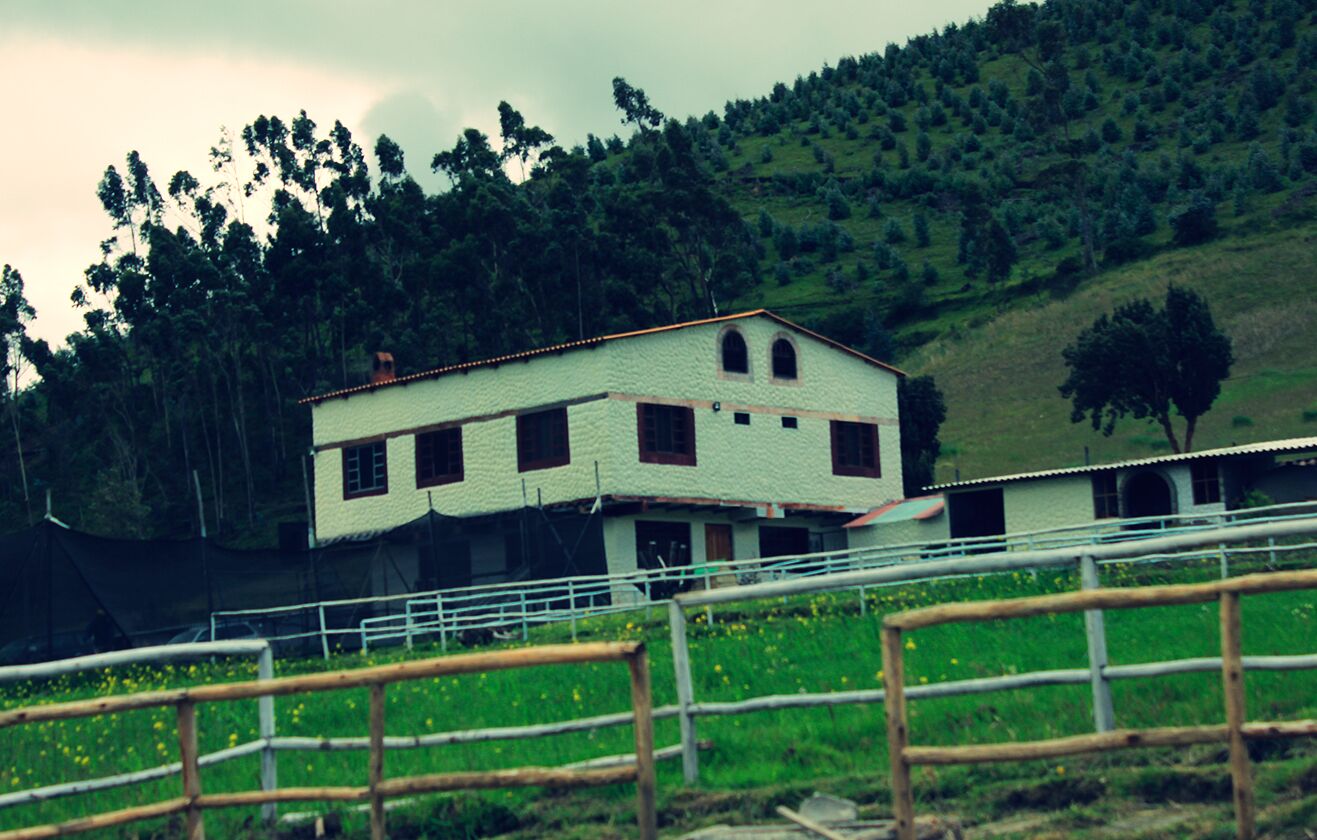 A two-story building with a white exterior and multiple windows, surrounded by a fenced area with a grassy field and trees.
