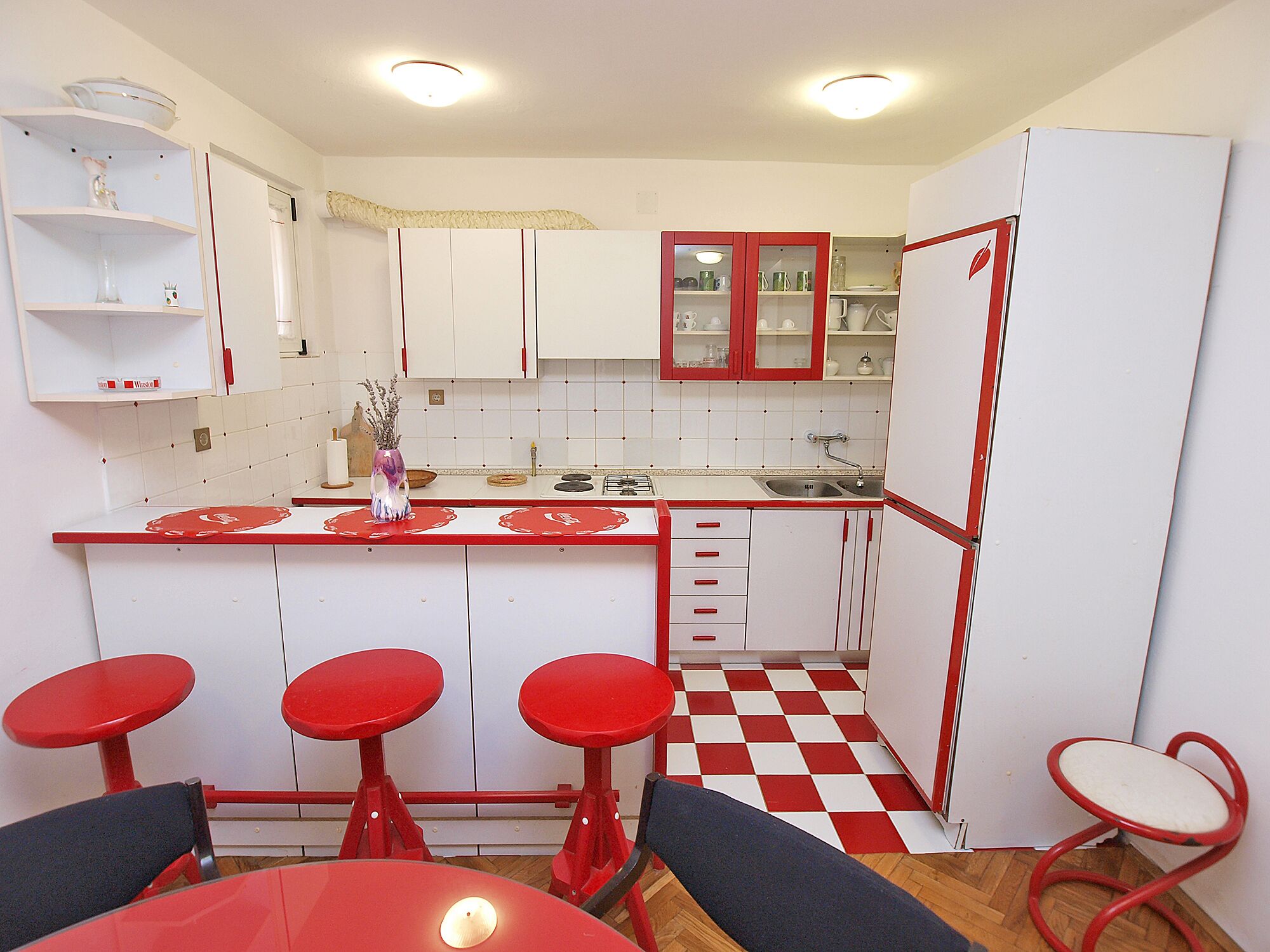 A compact kitchen with white cabinets, red accents, a checkered floor, and a bar area with stools.