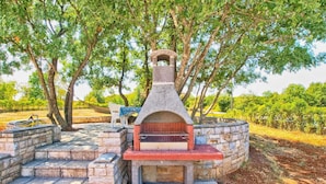 An outdoor stone barbecue area with steps leading up to it, surrounded by trees and a paved walkway.