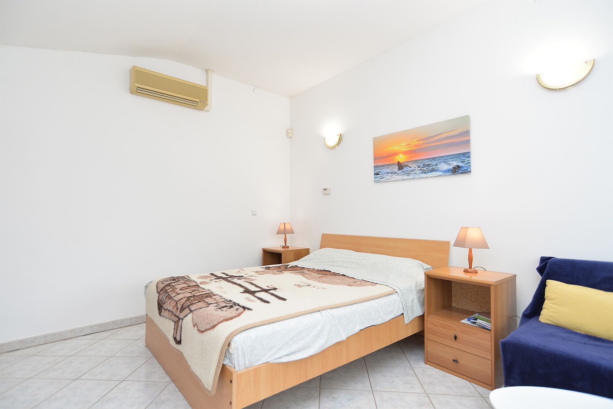 Une chambre à coucher comprenant un lit, des tables de chevet, une lampe, un climatiseur fixé au mur et un tableau représentant un coucher de soleil.