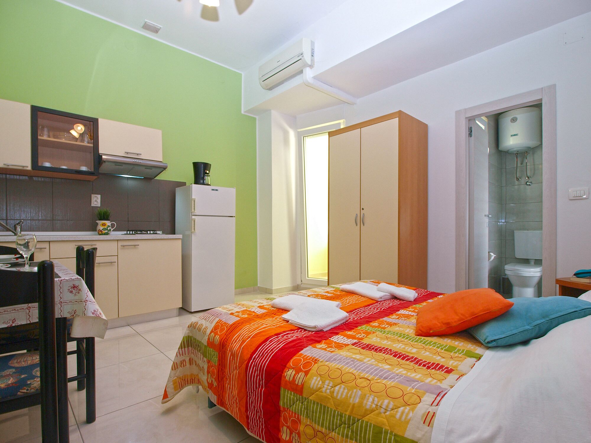 A compact bedroom with a bed, a small kitchenette, a wardrobe, and a bathroom.