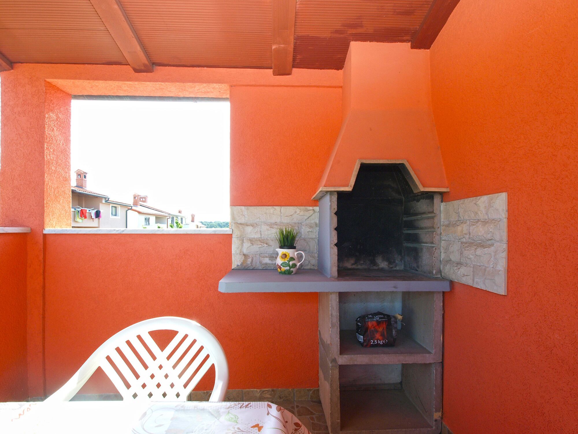 A small outdoor seating area with a stone fireplace, a white plastic chair, and a window overlooking a residential area.
