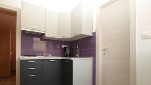 Une cuisine de taille réduite, avec des meubles de couleur sombre, une cuisinière à gaz, une machine à café et des murs recouverts de carreaux violets.