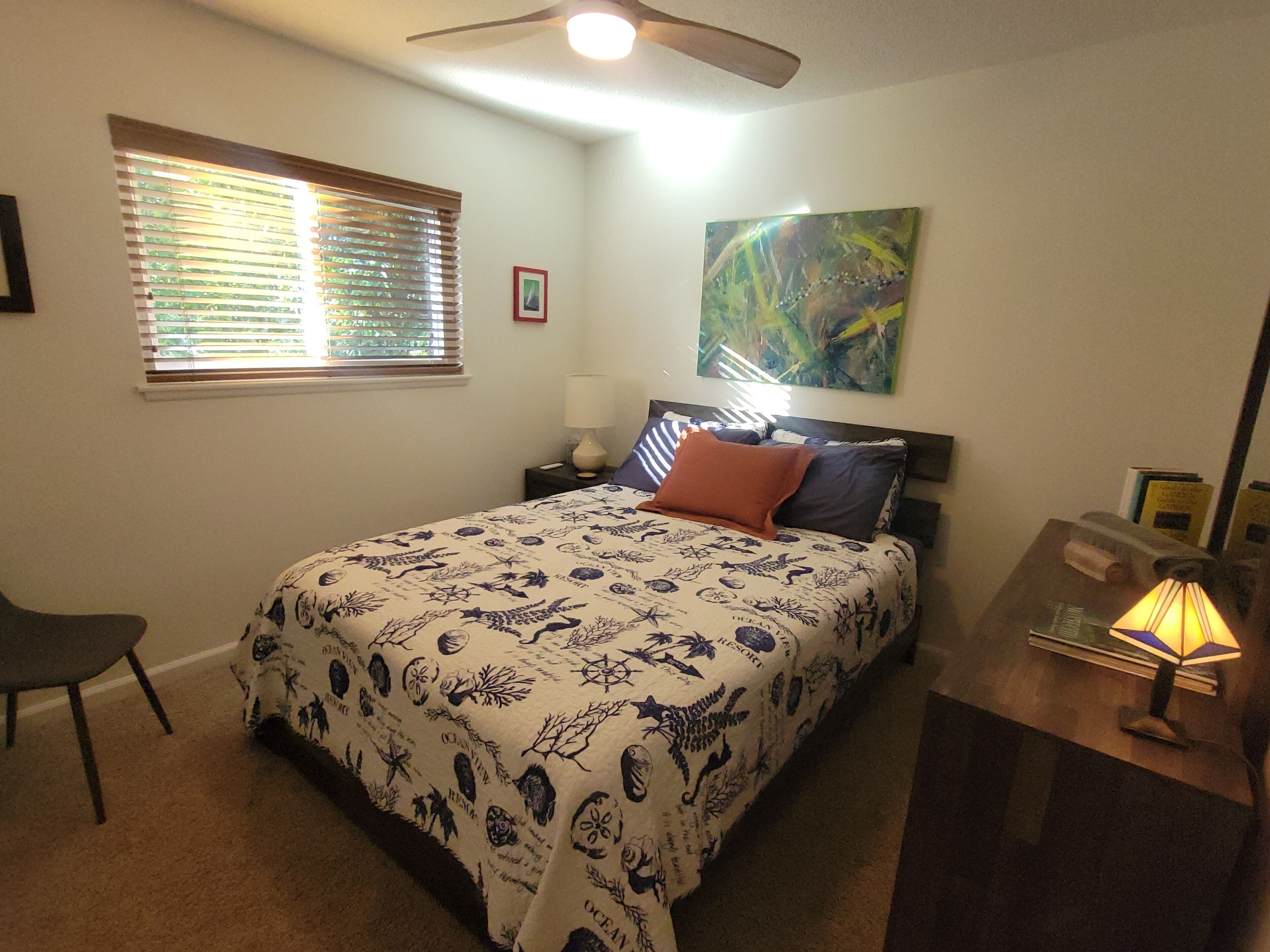 Ein Schlafzimmer mit Bett, Schreibtisch, Stuhl, Deckenventilator, Fenster mit Jalousien und einem Bild an der Wand.