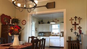 A dining area with a wooden table, chairs, and a vase of flowers. There is a kitchen in the background with white cabinets and a hanging light fixture.