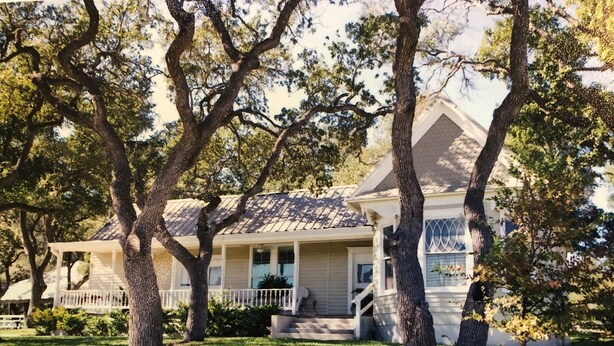 A two-story house with a porch, surrounded by trees and a stone retaining wall.