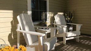 Two white Adirondack chairs on a porch with a table and a bottle of wine.
