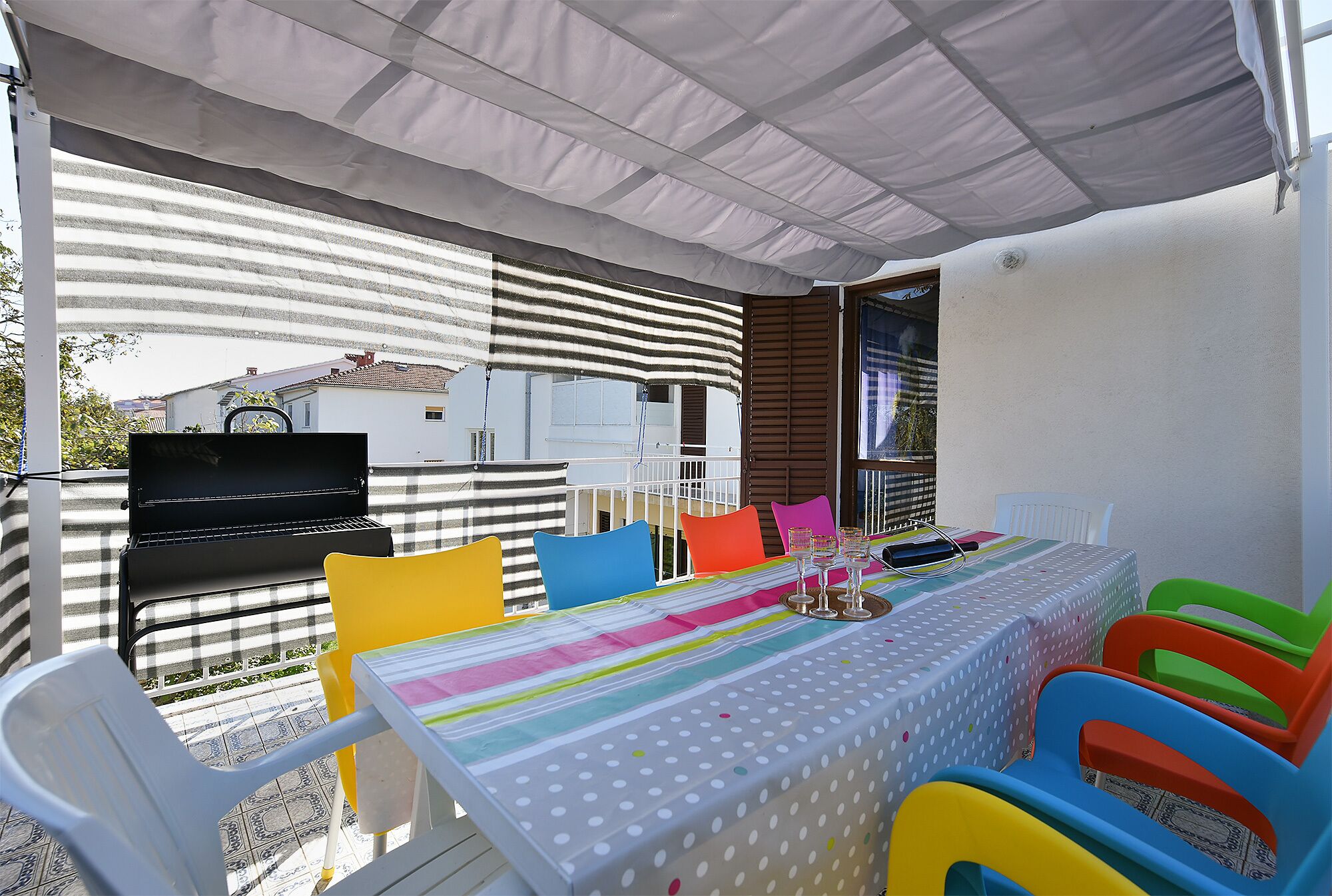 An outdoor patio with a striped awning, a grill, colorful chairs, and a table with a polka dot tablecloth.