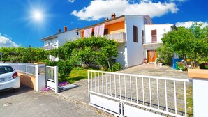 A two-story house with a white facade, a balcony with a railing, and a driveway with a white gate.