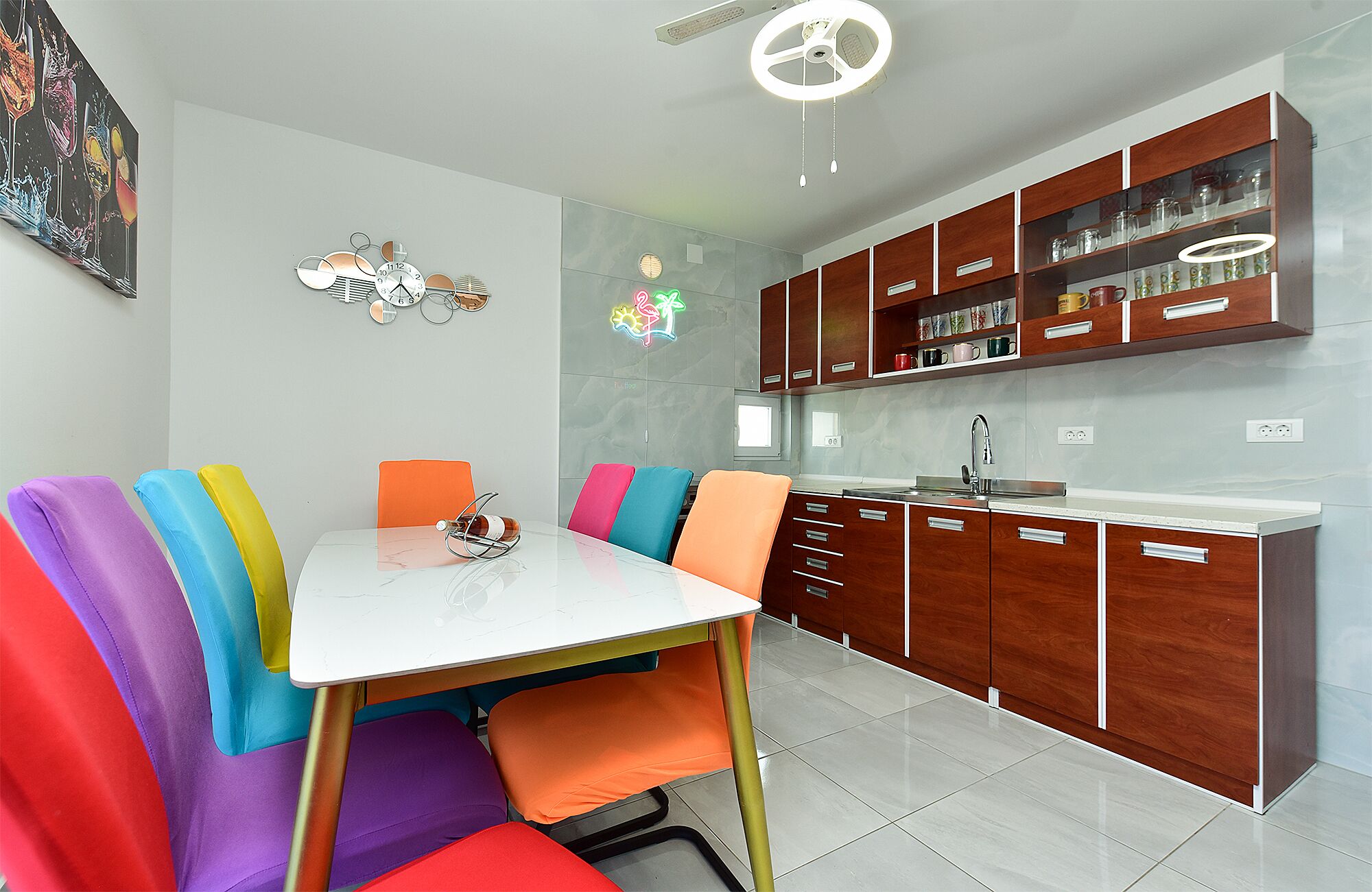 A modern kitchen with wooden cabinets, a white countertop, and a dining area with colorful chairs.