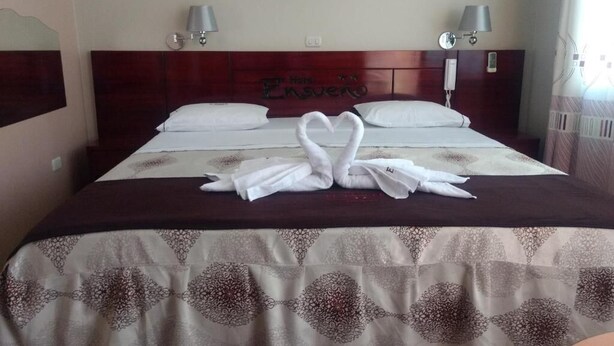 Habitación de hotel con cama, dos lámparas de noche, cabecera con el nombre del hotel y una decoración de toallas sobre la cama.