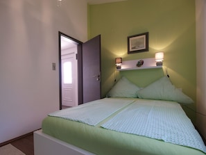 A bedroom with a bed, a nightstand, a wall-mounted lamp, and a door leading to another room.