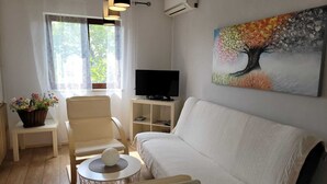 A living room with a white sofa, a chair, a small round table with a decorative item, a television on a stand, and a painting of a tree on the wall.