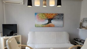 A modern living room with a white sofa, two white armchairs, a small white side table, a television, and a painting of a tree.